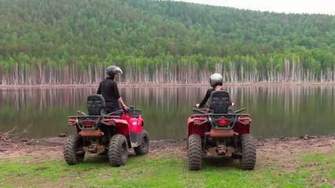 Aerial shot of couple in helmets sitting on red quad bikes by the lake in picturesque area