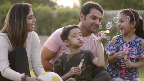 Happy family including mother, father, and two kids are blowing soap bubbles and enjoying the summer holidays in the garden or park. Smiling parents and children are spending leisure time together.
