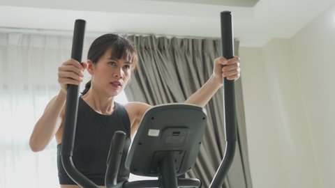 Asian woman working out on cycling machine at home. The sporty girl exercise on training equipment feeling tired and serious workout for her health at house during the COVID pandemic and citylock down Vídeo Stock