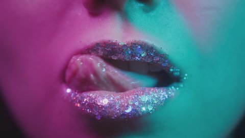Female lips in neon ultraviolet light. Fashionable neon light, euphoria. Sexy woman licks her teeth with her tongue. Shiny lips in a nightclub. Pink-blue-green color and new light. Metallic lipstick.