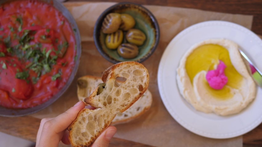 Brunch specialty in vegan restaurant. Top view of knife spreading hummus on no yeast bread with tomato dip and olives on side Royalty-Free Stock Footage #1061695987