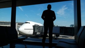Passenger stand against lounge window with arms a-kimbo. Passenger look out to large airliner, turn head aside, silhouetted shot. Guy wear smart casual suit and jeans - Powered by Shutterstock - Get 15% off with code: PIKWIZARD15