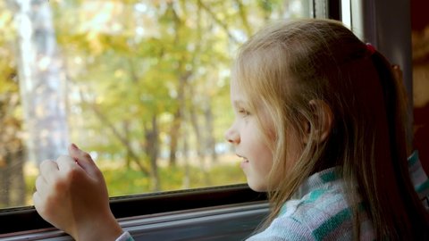 Cute little child girl smiles and looks out window of train in carriage during journey. Yellow trees and leaves background. Freedom concept, enjoy autumnal nature
