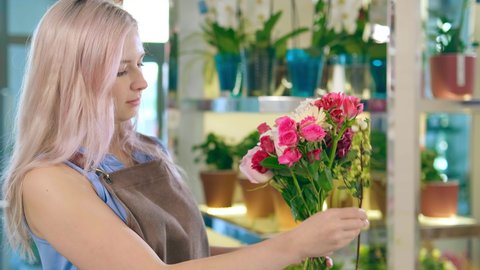 Nice blonde woman in light brown apron holds small flower stalks in hands making tender pink bouquet close view slow motion