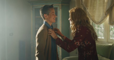 Cinematic shot of an young carefree happy smiling homosexual blond hair female gay is helping buttoning a shirt to her transgender man and giving affective kiss as sign of forever love at home.
