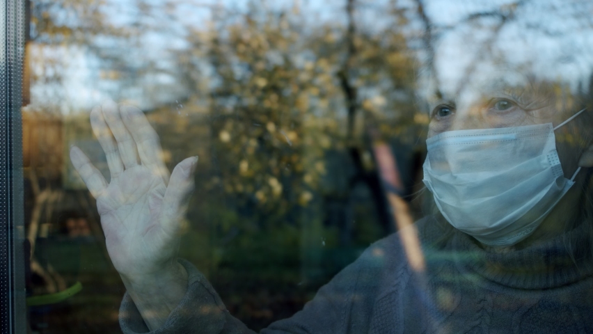 Portrait in the window of an elderly woman in a medical protective mask who put her hand on the glass and waiting for medics or relatives to help her | Shutterstock HD Video #1061700256