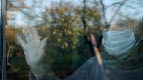 portrait in the window of an elderly woman in a medical protective mask who put her hand on the glass and waiting for medics or relatives to help her