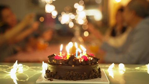 Chocolate cake with candle stick with light prepared on table for children's birthday party. People in family playing sparkler in background.