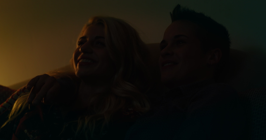 Cinematic shot of young carefree happy smiling homosexual female gay and transgender man couple in love is having fun watching television while sitting on a sofa in living room at home at night. Royalty-Free Stock Footage #1061700853
