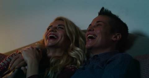 Cinematic shot of young carefree happy smiling homosexual female gay and transgender man couple in love is having fun watching television while sitting on a sofa in living room at home at night.
