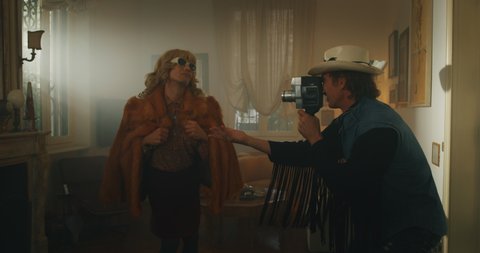 Cinematic shot of an young homosexual male gay is filming with vintage camera his carefree happy smiling transgender woman partner with sunglasses and wig while is having fun and dancing crazy at home