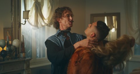 Cinematic shot of carefree happy smiling homosexual male gay dressed as cowboy is taking off a wig of his transgender woman partner and giving passionate kiss as sign of forever love at home.