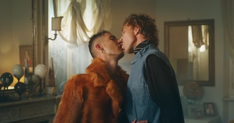 Cinematic shot of carefree happy smiling homosexual male gay dressed as cowboy is taking off a wig of his transgender woman partner and giving passion kiss as sign of forever love at home.