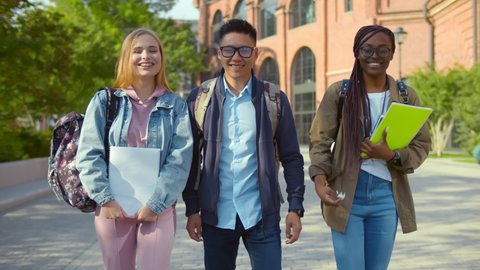 Happy multiracial students outdoor walking and talking to each other. Portrait of cheerful diverse college friends with books and backpack walking together at campus after classes