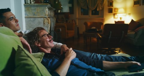 Cinematic shot of carefree happy smiling homosexual male gay and transgender woman couple in love is having fun watching television while lying with comfort on a sofa in living room at home at night.