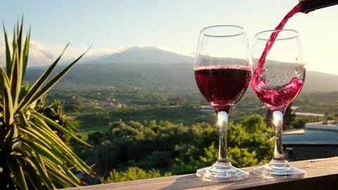 Pouring red wine from a bottle into two glasses on the vineyards in Sicily. Sommelier pouring red wine in glass with Etna volcano at background. Close-up of filling wine glass with red wine