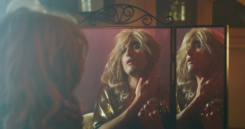 Cinematic shot of an young carefree homosexual male gay is putting on wig and in a mirror while preparing in vanity room. Concept of gender expression, identity and diversity.