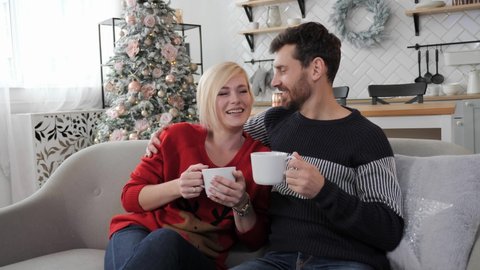 Festive mood, Christmas or New Year, a young couple sitting on the couch and drinking hot coffe. Christmas decor and modern white room