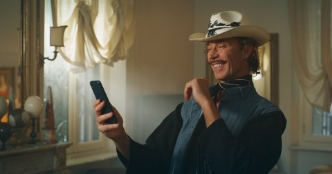 Cinematic shot of carefree middle aged happy man with moustache wearing cowboy hat is using a smartphone and smiling in camera in living room at home. Concept of technology, identity and diversity.