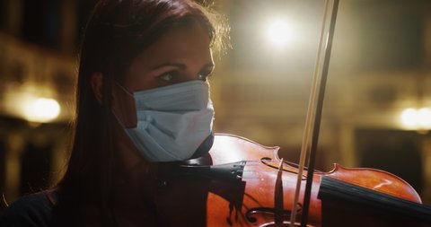 Cinematic close up shot of female violinist wearing protective medical mask is playing violin solo on a classic theatre stage with dramatic lighting during music concert.