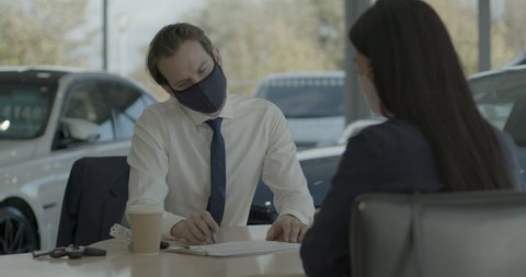 Car salesman dealer in auto showroom wearing face mask and customer signing contract and handing over key