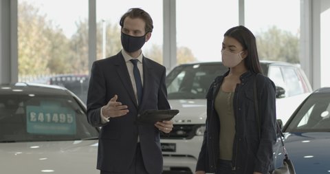 Car sales person talking to customer wearing protective face mask in car dealership showroom and looking at digital tablet