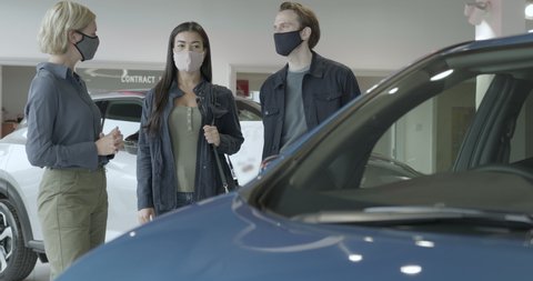 Couple with saleswoman looking at electric car in automotive showroom wearing protective face mask