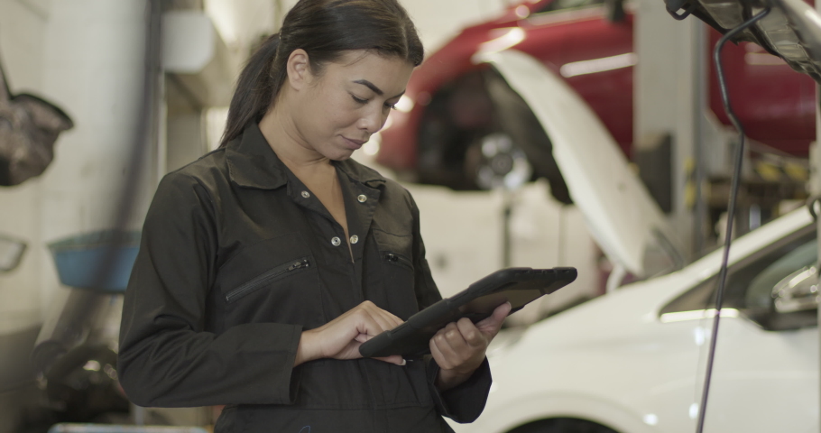 Female car mechanic looking under vehicle bonnet using digital tablet in automotive repair workshop