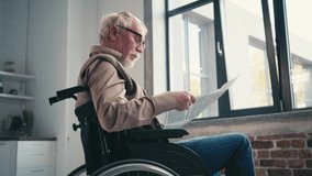 Side view of senior man in wheelchair reading newspaper near window at home