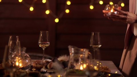Just before the celebration: woman hand's set a candle on Christmas decorated table with crystal glasses, wine bottle, candle and Christmas lights on the back