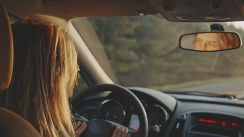 Rear view of happy young blonde in stylish sunglasses dancing to the music while driving her car at sunset. Pretty woman listening to enegetic music and smiling. Concept of joy and happiness