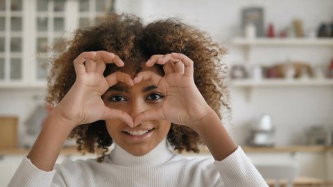 An attractive African American girl shows a heart gesture looks at the camera and smiles, a beautiful mixed race girl with curly lush hair looks at the camera and shows a heart on the eve of Valentine