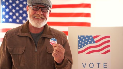 American retired masked to vote in 2020 US elections in the voting booth. An elderly man with glasses and a gray beard votes at a polling station holds a sticker in his hand and smiles proudly