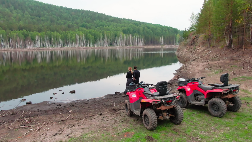 Drone shot of couple standing together by the lake and enjoying scenery after driving red quad bikes