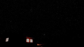 Small cabin in the mountains with light shining out the windows and a starry night sky in the background. Time lapse video.