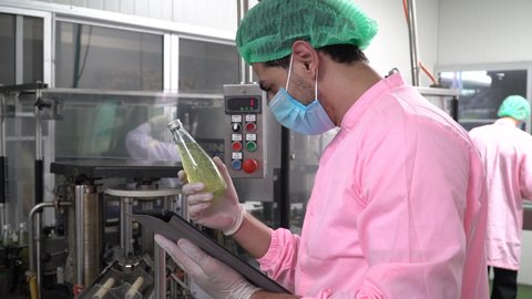 food scientist worker wearing hairnet and face mask checking quality control of juice bottle with tablet computer  in industrial water factory. inspection in line production beverage