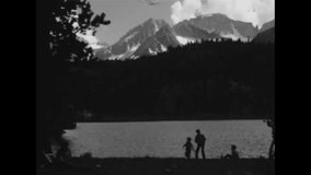 CIRCA 1960s - Some young kids play on a grassy area next to a big lake in the Sierra Mountains, as their parents set up camp and make a fire.