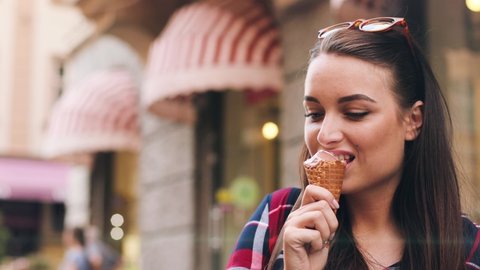 Beautiful young woman standing on street and eats ice cream in waffle cup. Attractive girl with brown hair in plaid shirt looks at camera and smiles.