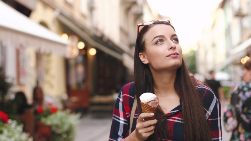 Beautiful young woman walks down the main street and eats ice cream in waffle cup. Attractive girl with brown hair in plaid shirt enjoys walk eating dessert.