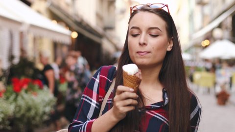 Beautiful young woman walks down the main street and eats ice cream in waffle cup. Attractive girl with brown hair in plaid shirt enjoys walk eating dessert.