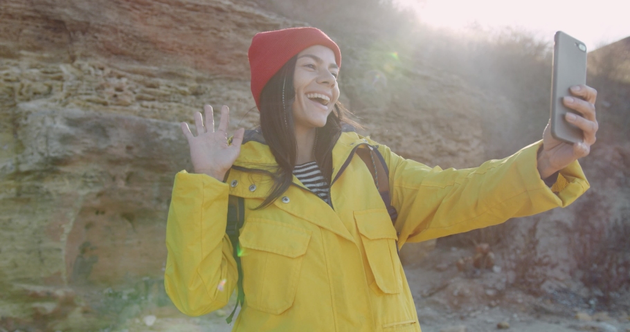 Beautiful female tourist in yellow jacket and red hat standing on background of large stones in sunlight and talking on video call using mobile phone.