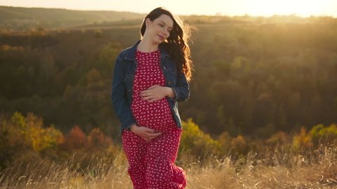 Pregnant woman looks at her belly and touches it with her hands. Healthy pregnancy. Autumn sunset, outdoors, slow motion.