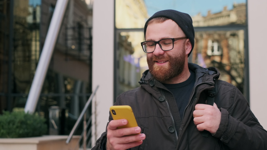Crop view of bearded man in glasses looking at phone screen and laughing while walking at street. Good looking guy scrolling social media news feed while using smartphone | Shutterstock HD Video #1061741410