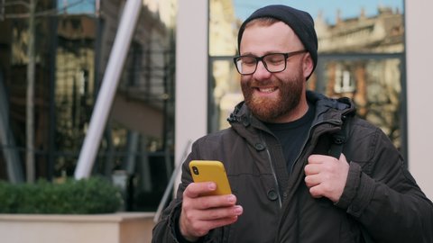 Crop view of bearded man in glasses looking at phone screen and laughing while walking at street. Good looking guy scrolling social media news feed while using smartphone
