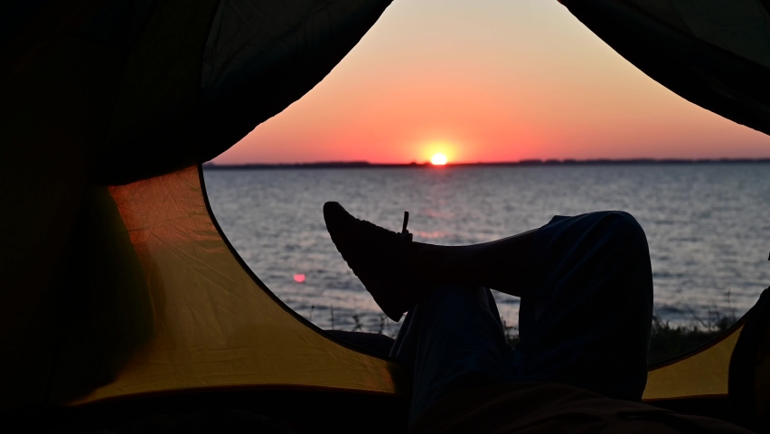 View of female legs in a tourist tent at sunset on the beach. The woman is hiking and camped on the riverbank. Royalty-Free Stock Footage #1061741656