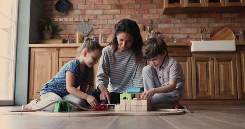 Loving young mother sit on warm floor playing with little son and daughter use wooden blocks and toy rail road having fun in modern kitchen. Communication, leisure playtime together with kids concept