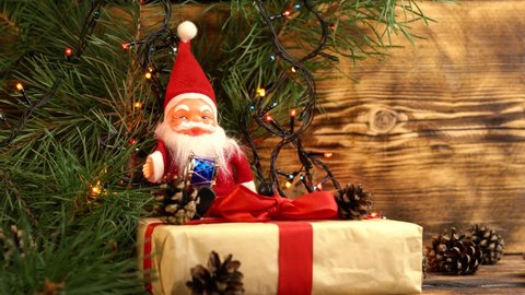 santa claus plush toy with a big white beard sits on a Christmas present with a red bow near the spruce branches with a garland and small cones on a wooden strom background