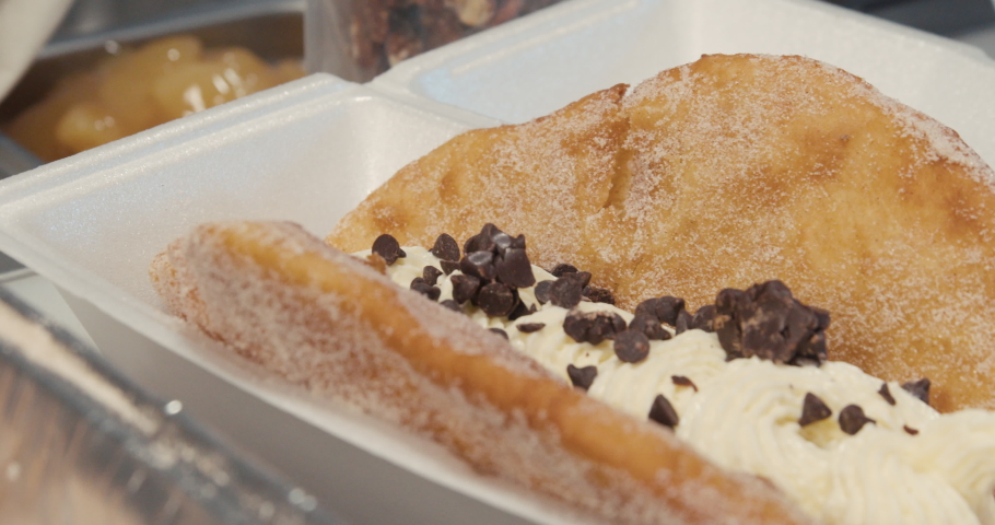 Sprinkling chocolate chips on a fried ice cream dessert. A gloved food truck worker sprinkles chocolate chunks on a decadent, sweet dessert with a crunchy fried shell, ice cream and whipped cream. Royalty-Free Stock Footage #1061744281