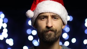 Young bearded European man wearing Santa hat and getting angry and stroking his beard. an angry European man in a Santa hat scratches his beard on a bokeh background. video close-up.