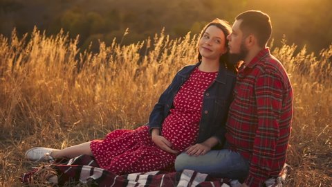 Happy couple of expectant parents, a pregnant woman and her husband are full of love. Picnic in nature at sunset. The sun hits the lens. Slow motion.